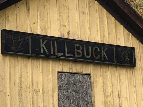 Sign on a yellow building displaying distances to Columbus (67 miles) and Hudson (66 miles), with "KILLBUCK" prominently featured.