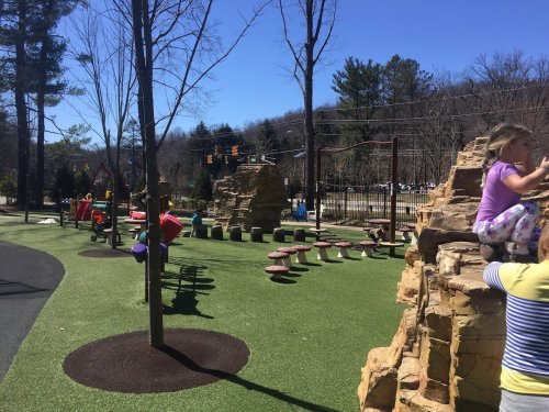 A sunny playground with climbing rocks, colorful equipment, and children playing on green turf surrounded by trees.