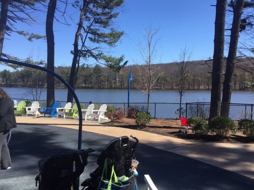 A serene lakeside view with chairs by the water, surrounded by trees and a playground area in the foreground.