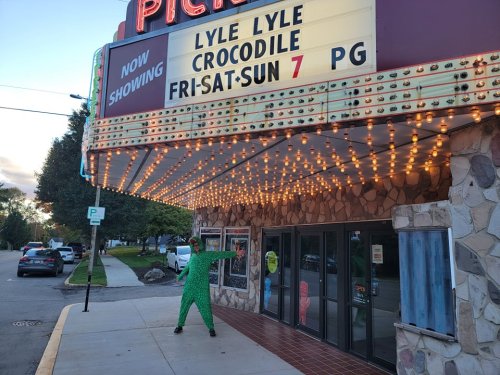 A person in a green crocodile costume poses in front of a theater marquee showing "Lyle Lyle Crocodile."