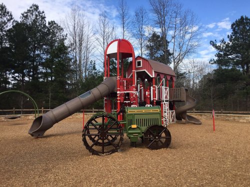 A colorful playground featuring a barn-themed structure, slides, and a toy tractor in a wooded area.