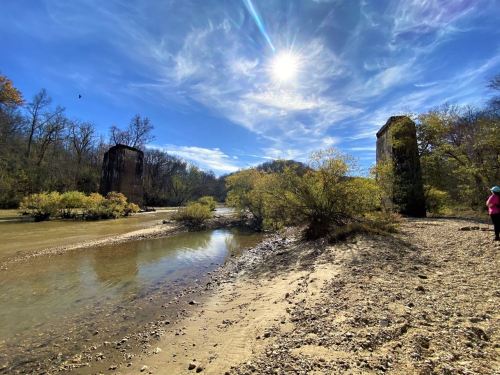 Ruins of tall structures by a riverbank, surrounded by trees and a bright blue sky with scattered clouds.