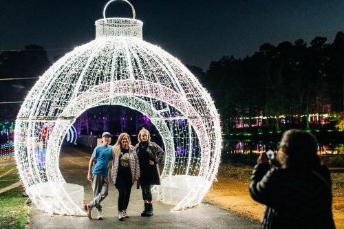 Three people pose inside a large illuminated ornament structure at a festive light display, with colorful lights in the background.
