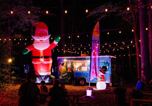 A festive scene with a large inflatable Santa, an ice cream truck, and string lights in a wooded area at night.