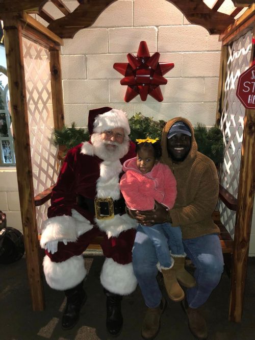 A man and a young girl sit on a bench with Santa Claus, smiling in a festive setting.