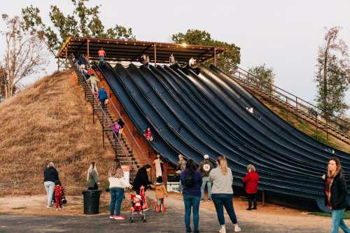 A large slide with people climbing stairs and sliding down, surrounded by a grassy area and trees at dusk.