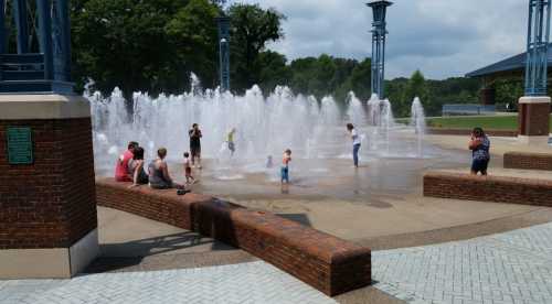 Children and adults play in a splash pad fountain on a sunny day, surrounded by greenery and a park setting.