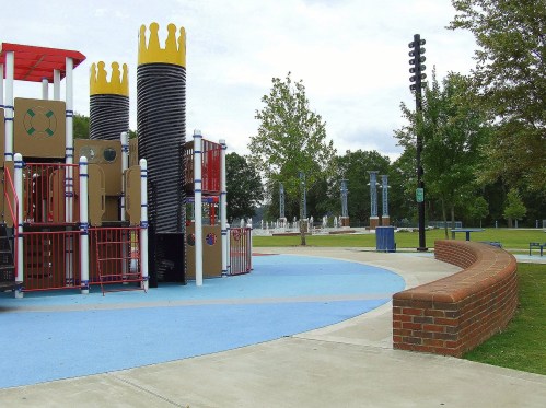 Colorful playground equipment with towers and a curved brick wall, set in a park with trees and a water feature in the background.