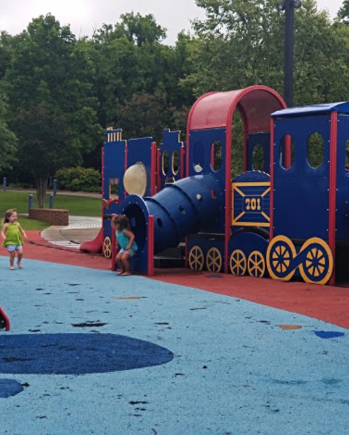 Children play on a colorful train-themed playground surrounded by green trees and a blue sky.