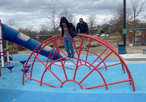 A child in a black hoodie balances on a red climbing structure at a playground, while an adult watches nearby.