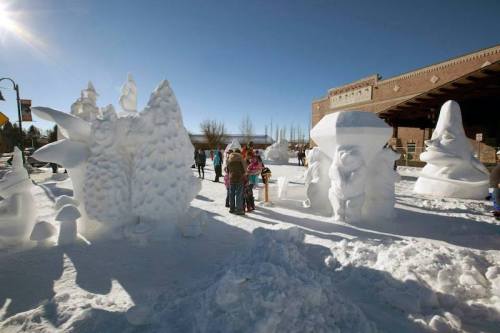 A snowy scene with large ice sculptures and people admiring them under a clear blue sky.