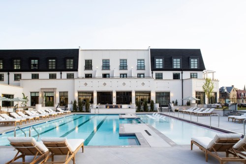 A modern hotel with a large outdoor pool and lounge chairs, surrounded by greenery and a clear blue sky.