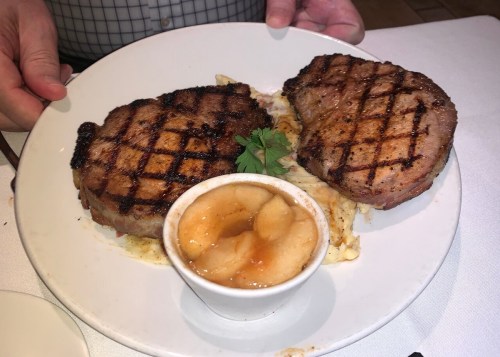 A plate with two grilled pork chops on a bed of mashed potatoes, accompanied by a small bowl of applesauce.
