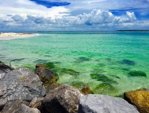 A serene beach scene with clear turquoise water, rocky shore, and a cloudy blue sky in the background.