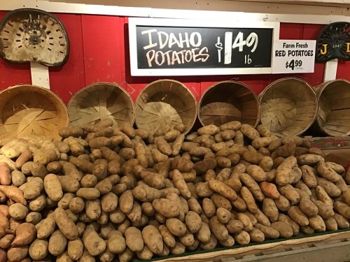 A display of Idaho potatoes stacked on a table, with baskets in the background and price signs for potatoes.