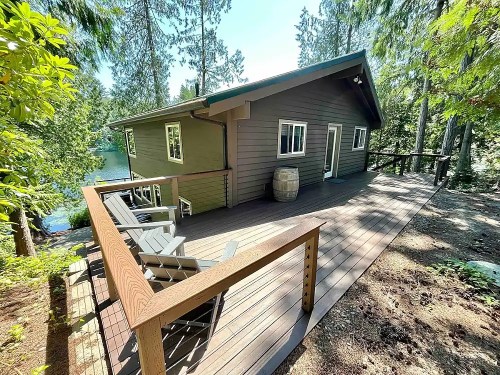 A cozy cabin with a deck overlooking a serene lake, surrounded by tall trees and greenery.