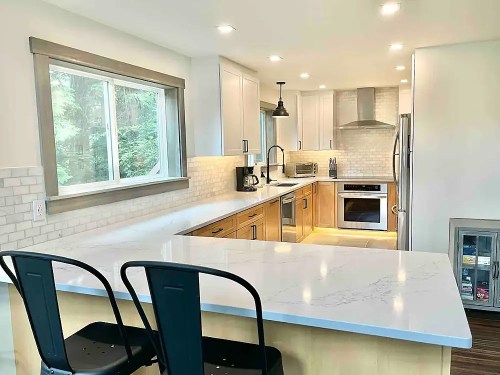 Modern kitchen with light wood cabinets, white countertops, stainless steel appliances, and a large window.
