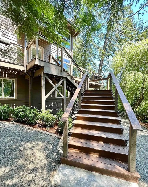 A wooden staircase leads up to a house surrounded by trees and greenery on a sunny day.