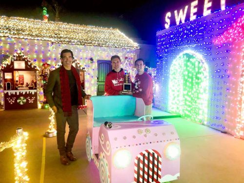 Three people stand in front of a brightly lit holiday display, featuring colorful decorations and a festive atmosphere.