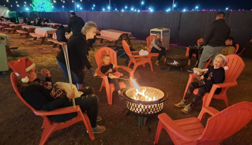A cozy outdoor scene with people in red chairs around a fire pit, enjoying snacks and holiday decorations.