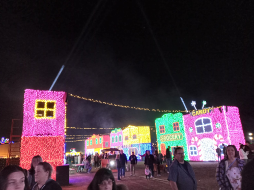 Colorful holiday lights illuminate a festive scene with candy-themed buildings and crowds enjoying the display at night.