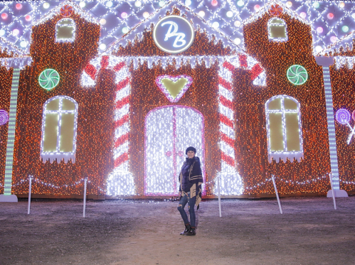 A person stands in front of a brightly lit, festive gingerbread house display adorned with colorful candy decorations.