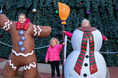 Two children pose with large gingerbread and snowman decorations, smiling in front of a festive backdrop.