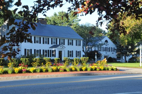 A charming white house with black shutters, surrounded by colorful flower beds and trees, along a quiet street.