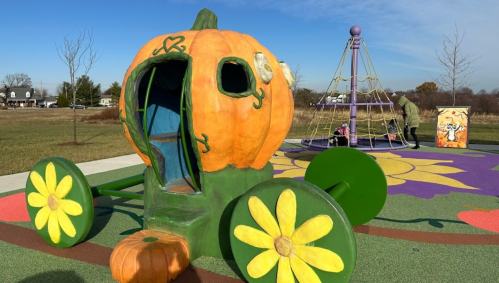 A colorful pumpkin-shaped play structure with flowers, set in a park on a sunny day. Children play nearby.