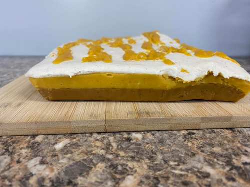 A layered dessert on a wooden board, featuring a yellow base and a white topping with orange drizzle.