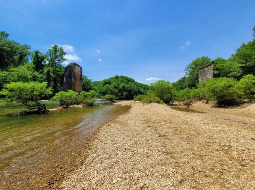 A serene riverbank scene with two tall, weathered structures surrounded by lush greenery and clear blue skies.