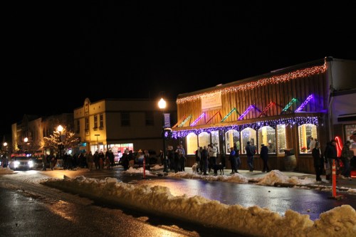 A festive street scene at night, with shops decorated in colorful lights and a crowd walking along the snowy sidewalk.