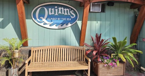 A wooden bench in front of a teal wall with a sign reading "Quinn's Almost by the Sea" and potted plants nearby.