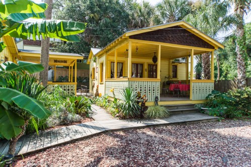 A charming yellow cottage with a porch, surrounded by lush greenery and tropical plants.