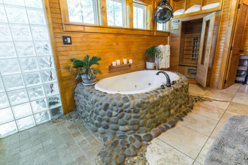 A luxurious bathroom featuring a stone-accented bathtub, large windows, and a sauna in a wooden setting.