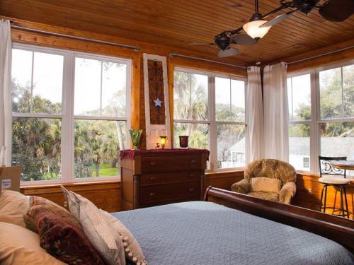 Cozy bedroom with wooden ceiling, large windows, a bed, dresser, and a comfortable chair, surrounded by greenery.