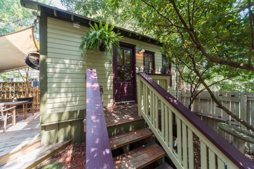 A small green cabin with a wooden ramp, surrounded by trees and outdoor seating areas.