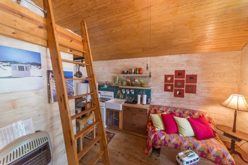 Cozy wooden interior with a loft, colorful sofa, kitchen area, and a wooden ladder leading to the upper level.