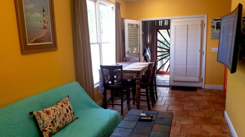 Cozy living space with yellow walls, a turquoise couch, dining table, and large windows letting in natural light.