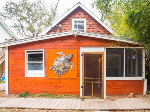 A bright orange house with a turtle mural, featuring a screened porch and wooden walkway surrounded by greenery.
