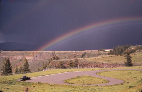 A winding road curves through green fields under a dramatic sky with a vibrant rainbow arching above.