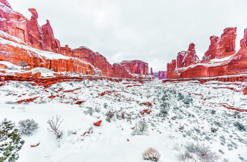 A snowy landscape featuring red rock formations and cliffs under a cloudy sky.