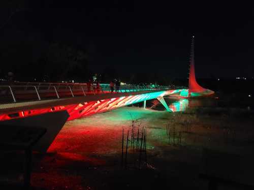 A brightly lit pedestrian bridge at night, featuring red and green lights, with people walking across it.