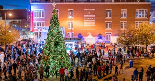 A festive crowd gathers around a large, decorated Christmas tree in a lively downtown area at night.