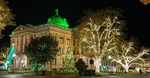 A beautifully lit historic building with green accents, surrounded by festive trees and holiday lights at night.