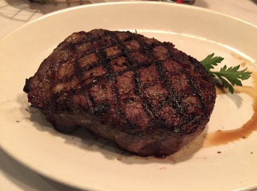 A perfectly grilled steak on a white plate, garnished with a sprig of parsley and drizzled with sauce.