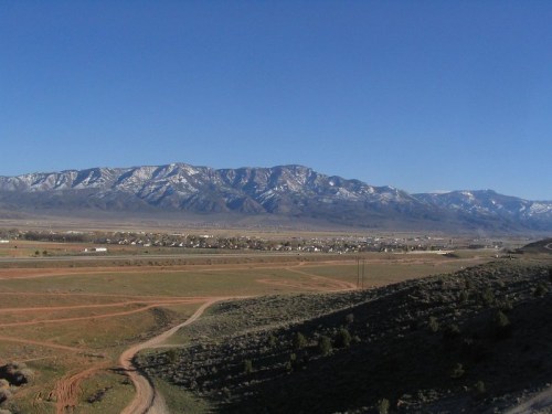 A panoramic view of snow-capped mountains over a valley with dirt roads and sparse vegetation under a clear blue sky.