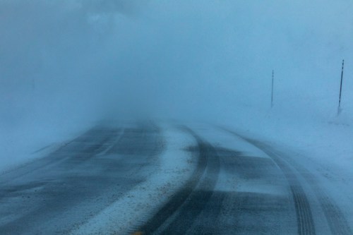 A snow-covered road disappears into thick fog, creating a sense of isolation and winter conditions.