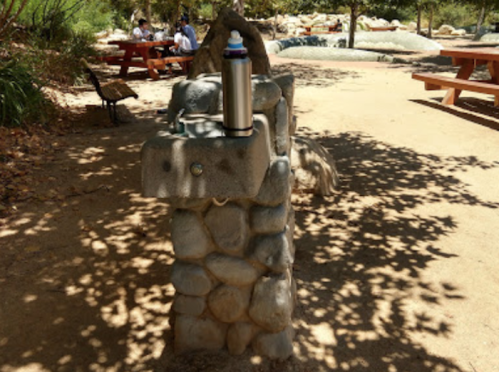 A stone water fountain with a metal water bottle on top, surrounded by trees and picnic tables in a park setting.