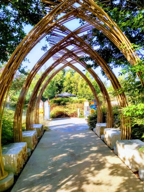 A serene garden pathway framed by arching bamboo structures, surrounded by lush greenery and colorful plants.